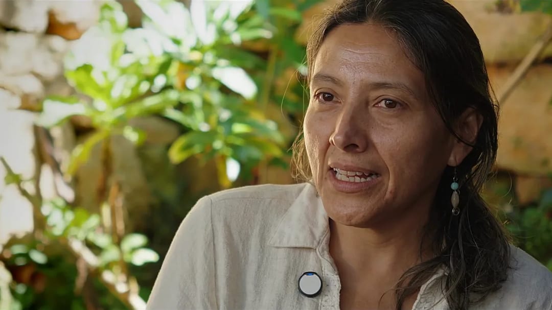Una mujer conversa sentada frente a un muro de piedra rodeada de vegetación