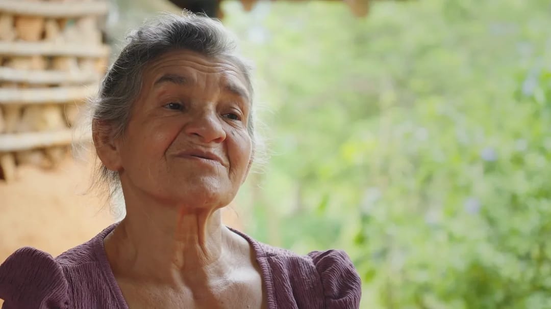 Una mujer campesina sonríe sentada frente a la vegetación de su finca