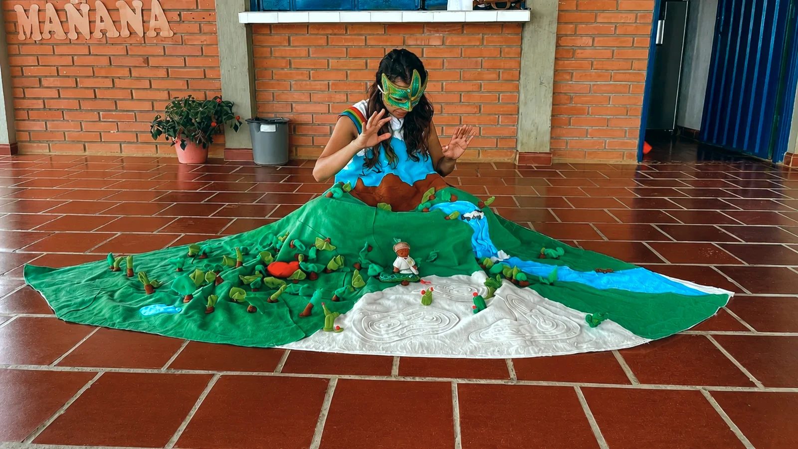 La Madre Tierra, con máscara de hojas y una falda que es un paisaje de bosque y río, sentada en el piso de una escuela