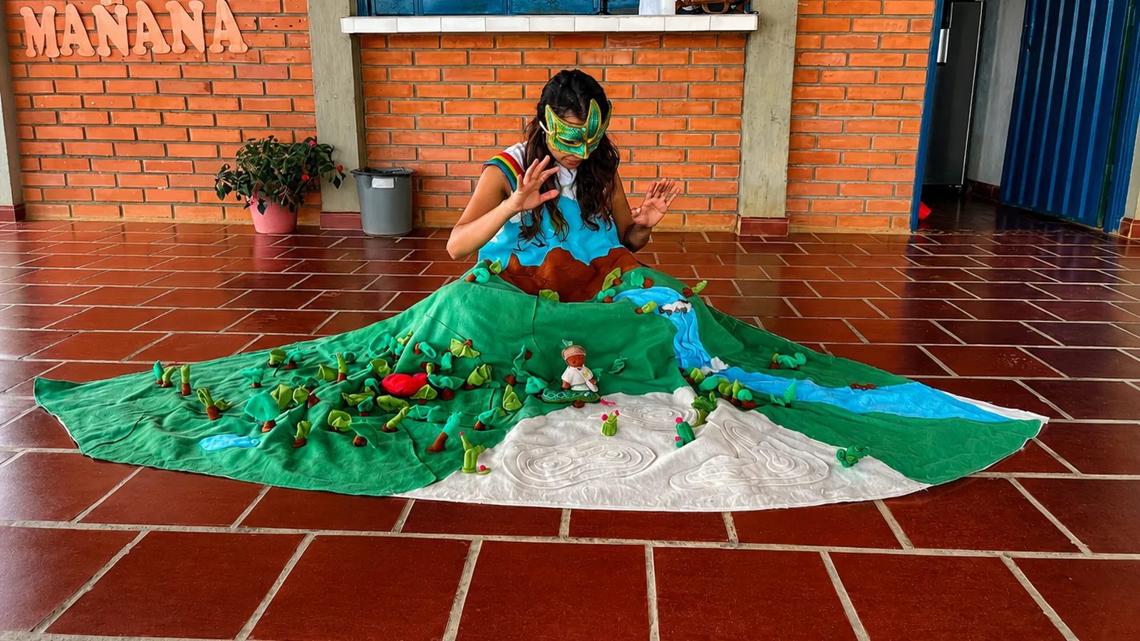 La Madre Tierra, con máscara de hojas y una falda que es un paisaje de bosque y río, sentada en el piso de una escuela